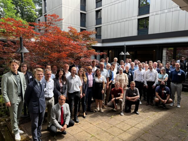 A large group of people standing and kneeling together for a casual business photo outside buildings at the Karlsruhe Institute of Technology, with a red-leaf tree in the foreground and green foliage in the background.