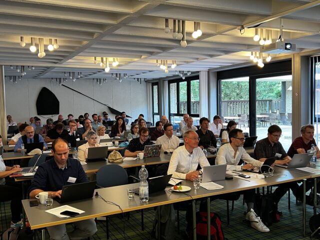 A large group of adults from seated at rows of tables in a conference room, many using laptops and facing toward the front of the room. There are large windows along one side of the room overlooking greenery outside. Laptops, water bottles and notebooks are visible on the tables.