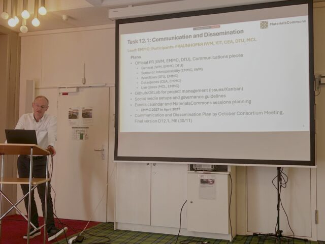 Gerhard Goldbeck stood leant against a lectern at the front of a meeting room. A large projection screen displays a slide titled “Communication and Dissemination”.